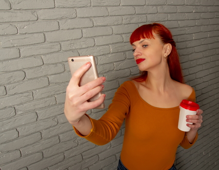 Young red-haired girl with a cup of coffee makes selfie with her smartphone on a background of gray brick wallの写真素材