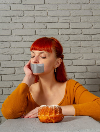 Conceptual image of a young red-haired girl on a diet sitting with her mouth taped with silver tape...