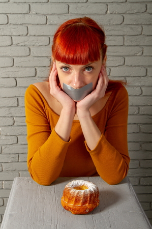 Conceptual image of a young red-haired girl on a diet sitting with her mouth taped with silver tape before appetizing pastriesの写真素材