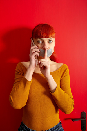 Conceptual picture. Young red-haired girl tries to talk on the phone with sealed with adhesive tape with her lips so that she keeps silenceの写真素材