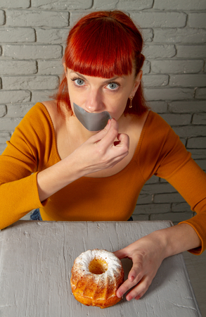 Conceptual image of a young red-haired girl on a diet sitting with her mouth taped with silver tape before appetizing pastriesの写真素材