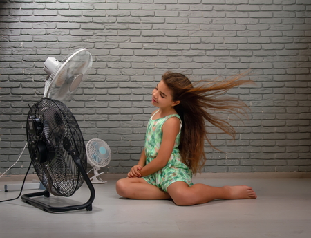 A little girl cools herself from the heat with the help of three different fans at once sitting on the floor of the roomの写真素材