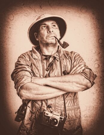 Adult Tourist in a tropical cork helmet and protective clothing smoking a pipe with his arms folded across his chest. Photo artificially aged as vintageの写真素材