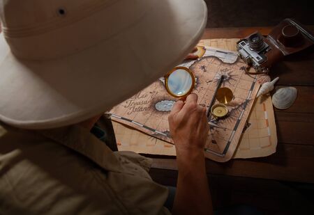 An adult explorer in a cork helmet over an old wooden table paves a route using old mapsの写真素材