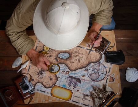 An adult explorer in a cork helmet over an old wooden table paves a route using old mapsの写真素材