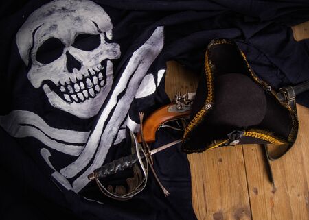 Classic pirate black felt captain's cocked hat with a pistol and a sword lying on a wooden floor next to the Jolly Roger flagの写真素材