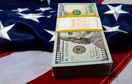 a pack with ten thousand dollars in hundred dollar bills lies against the backdrop of a starry striped American flagの写真素材