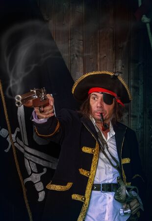 Adult pirate captain in a traditional costume shoots a pistol and holds a sword in his hands against the background of a jolly rogerの写真素材