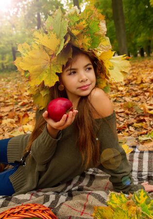 Little girl on a picnic in autumn park eats a ripe red apple sitting on fallen foliage in a headdress from maple leavesの写真素材