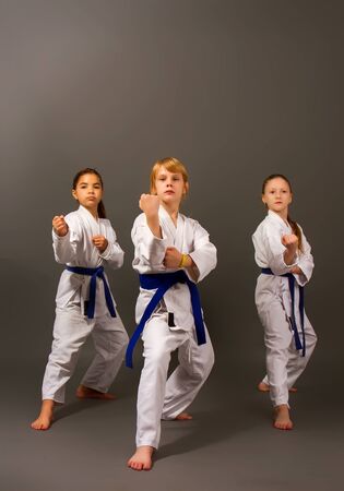 three little karate girls in a white kimono and blue belts perform a beautiful team kata on a dark backgroundの写真素材