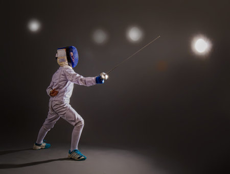Little boy in a white protective suit fencing mask and with a rapier exercises in fencing on a dark...