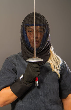 Young blonde girl in a fencing trainer dark uniform and black helmet mask stands with a rapier on a...