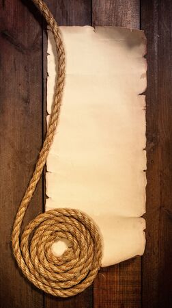 old sheet of parchment or paper lying on wooden boards and a coarse rope pulled into a nautical knot forming a frameの写真素材