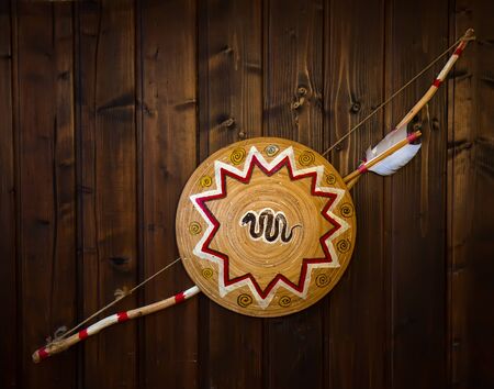 Native American weapons bow and arrow and patterned shield hang on a dark wooden wall.の写真素材