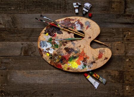 wooden classic palette smeared with multi-colored paints lying on a wooden background next to used brushes and tubes of paintの写真素材