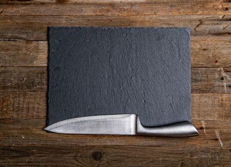 Stone slate flat black plate and sharp steel kitchen knife on a wooden backgroundの写真素材