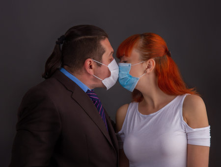 A man in a suit and a girl kiss on a dark background through protective medical masks.の写真素材