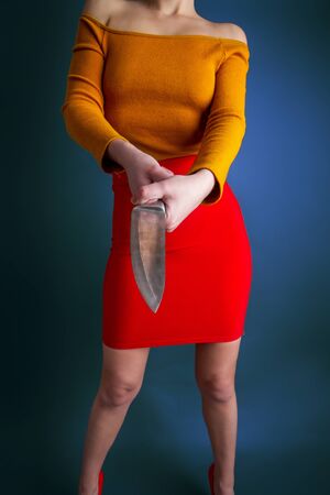 A young girl in a bright skirt and blouse holds in her hands a large steel kitchen knife on a dark backgroundの写真素材