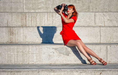 Young beautiful girl photographer in a bright red dress focuses on a model with a large digital cameraの写真素材
