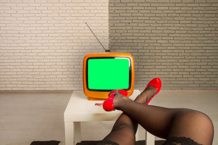 The girl sits in front of a small old TV on which the green screen is throwing her legs in bright red shoes on the table.の写真素材