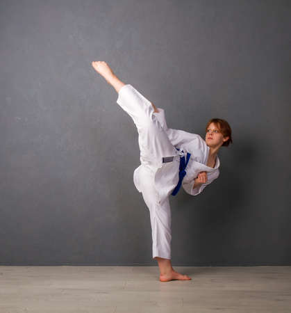 A young girl karateka in a white kimono and a blue belt trains and performs a set of exercises against a gray wallの写真素材