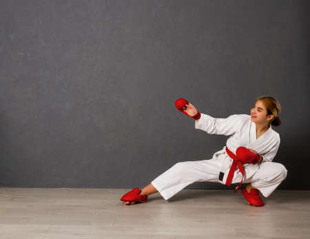 A young karateka girl in a white kimono and red competition outfit trains and performs a set of exercises against a gray wallの写真素材