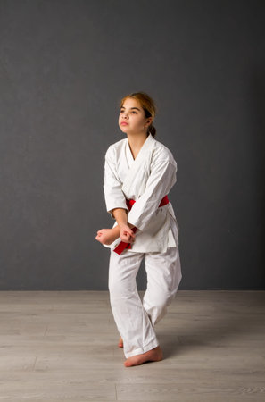 A young girl karateka in a white kimono and a red belt trains and performs a set of exercises against a gray wallの写真素材
