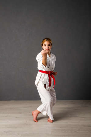 A young girl karateka in a white kimono and a red belt trains and performs a set of exercises against a gray wallの写真素材