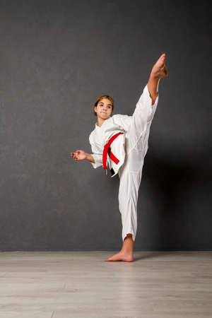 A young girl karateka in a white kimono and a red belt trains and performs a set of exercises against a gray wallの写真素材