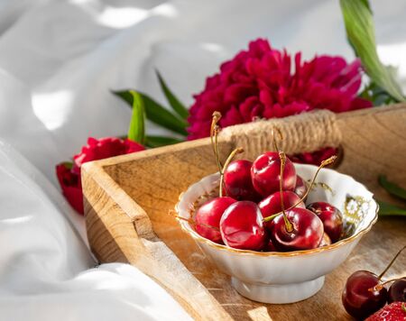 Peony and wooden rustic tray with bowls of strawberries and sweet cherry and cup of coffe on a white bedlinen. Morning mix of fruits, flowers an cup of cofee on a bedの写真素材