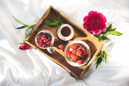 Peony and wooden rustic tray with bowls of strawberries and sweet cherry and cup of coffe on a white bedlinen. Morning mix of fruits, flowers an cup of cofee on a bedの写真素材