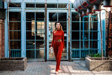 beautiful tall young girl model with long blond hair in an orange jumpsuit on the background of large windows with blue frames, goes and looks into the cameraの写真素材