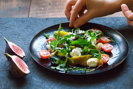Cooking salad on a black plate. Arugula, cherry tomatoes, curd cheese, dark grapes, figs, thyme. Human handの写真素材