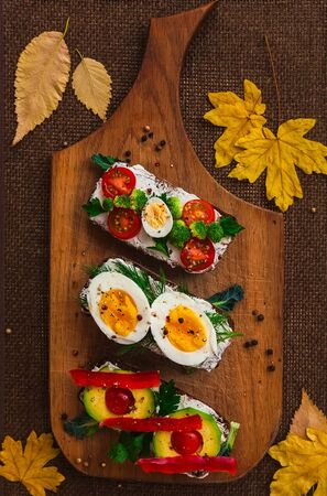 three bruschettas on dark bread with curd cheese, eggs, vegetables, spices and herbs on a wooden board, the background is decorated with yellow autumn leavesの写真素材