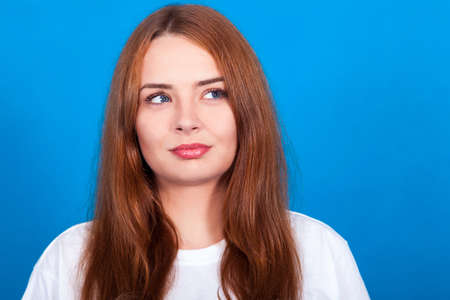 beautiful redhead girl with freckles on a blue background thinks, makes plans, sly look