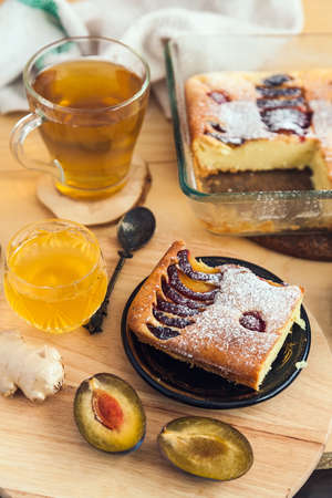 a square slice of plum pie on a round black antique plate, acacia honey, a vintage teaspoon and a cup of green teaの写真素材