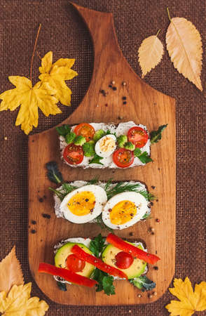 three bruschettas on dark bread with curd cheese, eggs, vegetables, spices and herbs on a wooden board, the background is decorated with yellow autumn leavesの写真素材