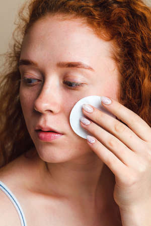 Teenage skin care concept. Young beautiful red-haired girl with long curly hair cleanses her face with a cotton pad. No makeup, no retouching. Natural beauty. Close-up shot in studioの写真素材