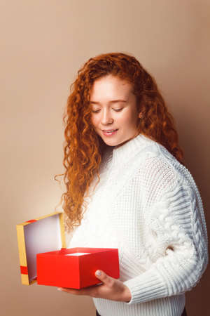 beautiful red-haired girl with long curly hair in a white knitted sweater opens a box with a gift and smilesの写真素材
