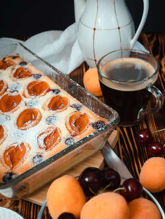 homemade apricot cake with curd dough, decorated with icing sugar and sweet cherry in a rectangular glass shape, a cup of black coffee and a white teapot on a dark wooden tableの写真素材