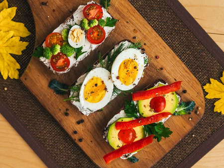 three bruschettas on dark bread with curd cheese, eggs, vegetables, spices and herbs on a wooden board, the background is decorated with yellow autumn leavesの写真素材