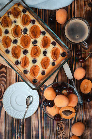 cottage cheese pie with apricots and cherries in a rectangular glass baking dish on a wooden table with a white plate and a cup of coffee, flat layの写真素材
