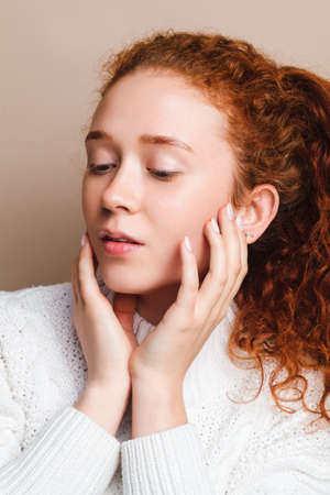 beautiful teenage girl with red curly hair, brown eyes and freckles in a white sweater looks at the camera. Shot in a studio on a beige background. Natural beauty conceptの写真素材