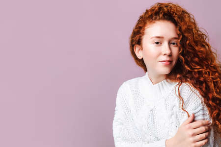 Cute smiling girl with long curly red hair in a white sweater in the studio. Copy space for your text on a pink background. Winter Skin Care Conceptの写真素材