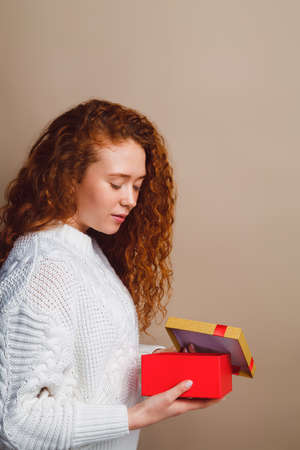 beautiful girl with long curly hair in a white sweater smiles and opens a red box with a golden lid - a gift. Shot in a studio on a beige background. Place for your text. Verticalの写真素材