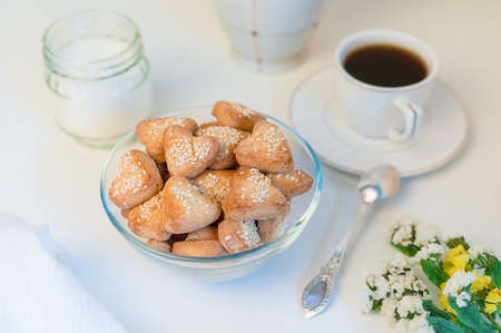 homemade heart shaped sesame shortbread cookies in a glass bowl, a cup of black coffee, sugar and wildflowers on a white table close upの写真素材
