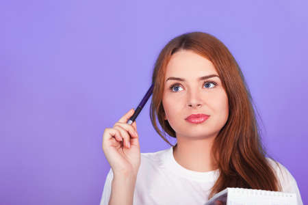 beautiful young red-haired girl with blue eyes in a white t-shirt thinks what to write in a...