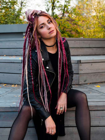 attractive young woman in black clothes with pink de-dreadlocks sits on wooden steps and looks at the cameraの写真素材