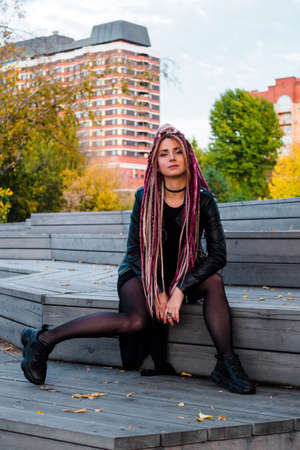 attractive young woman in black clothes with pink de-dreadlocks sits on wooden steps and looks at...
