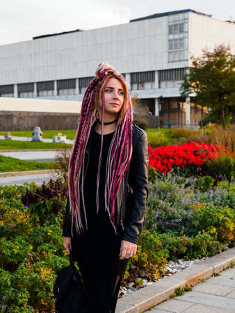 beautiful young woman with hairstyle - long pink and beige dreadlocks in black clothes walks in the...